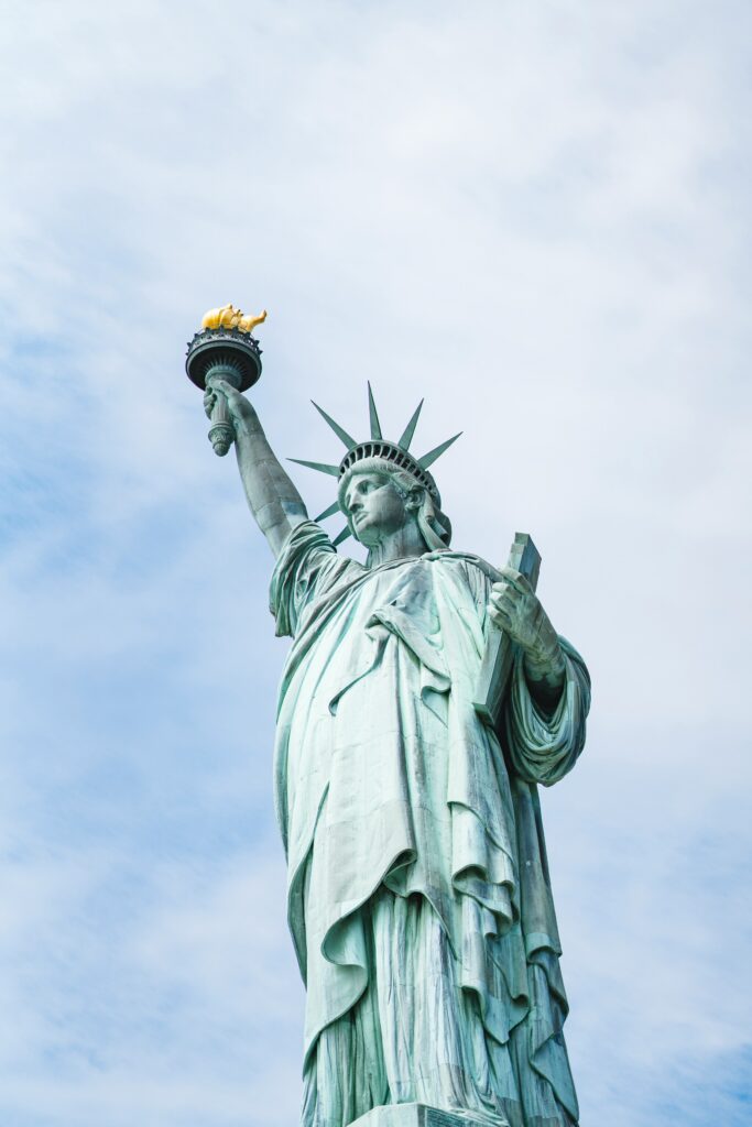Low-angle shot of the Statue of Liberty holding her golden torch high against a bright, lightly clouded blue sky.
